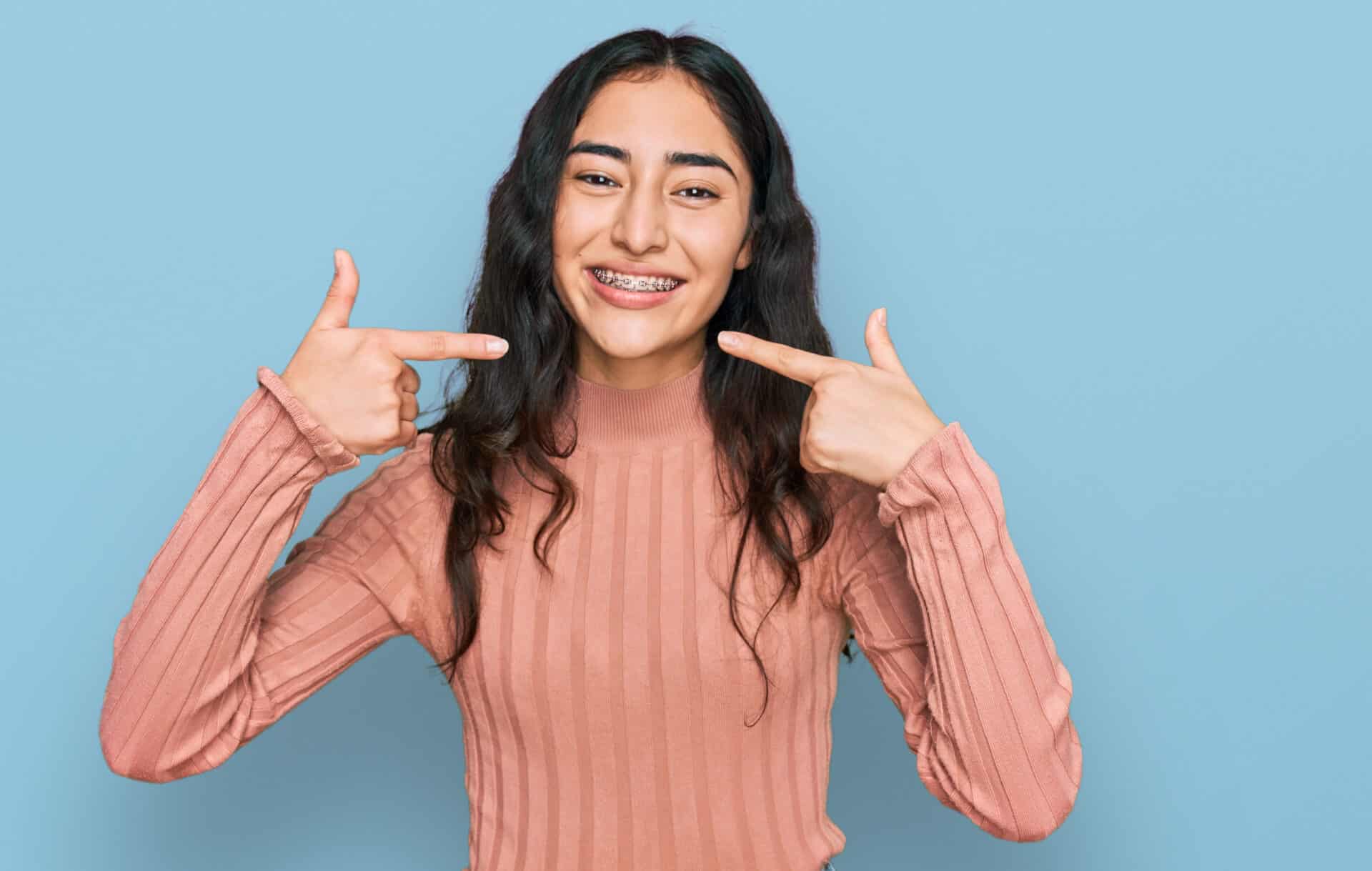 Hispanic teenager girl with dental braces wearing casual clothes smiling cheerful showing and pointing with fingers teeth and mouth. dental health concept.